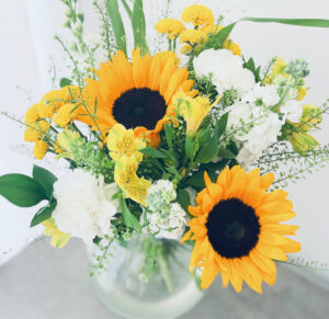 Sommarbukett med inslag av gula solrosor. Blommor i gult och vitt plus dekorationsgrönt. Buketten finns hos Made4y.se.